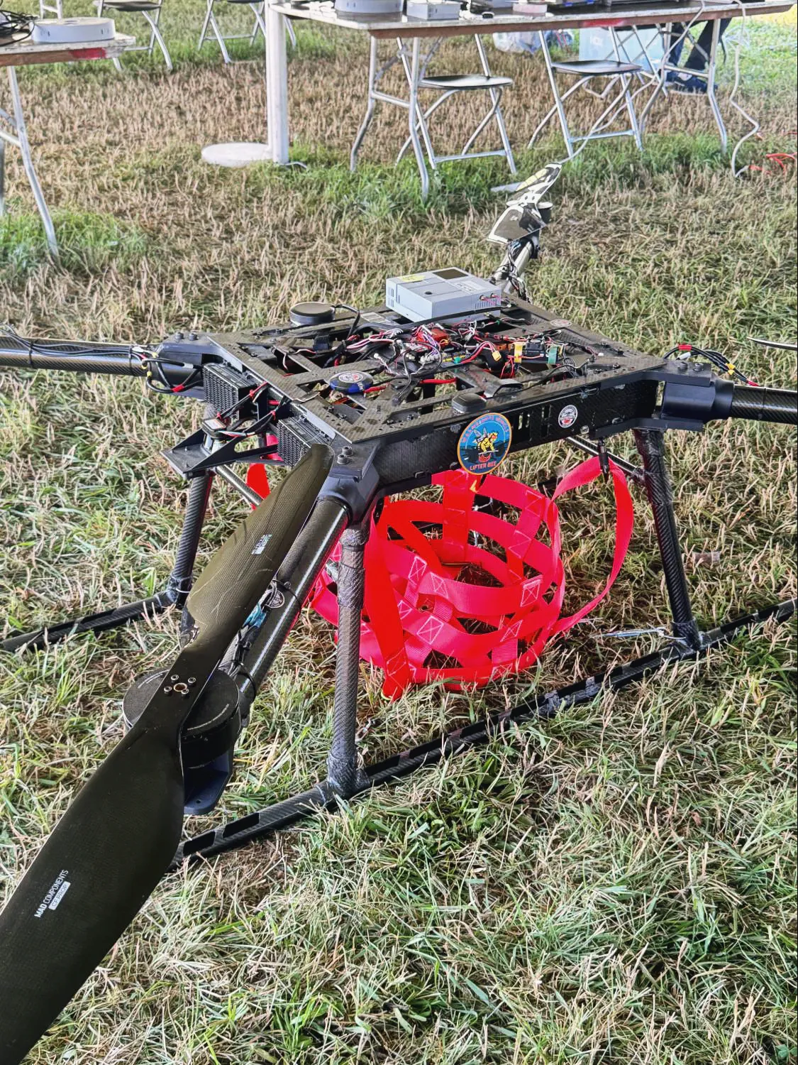 The MW-V1 airframe on grass under the tent, carbon arms and folded propellers around the centre plate, with the red cargo harness stowed underneath.