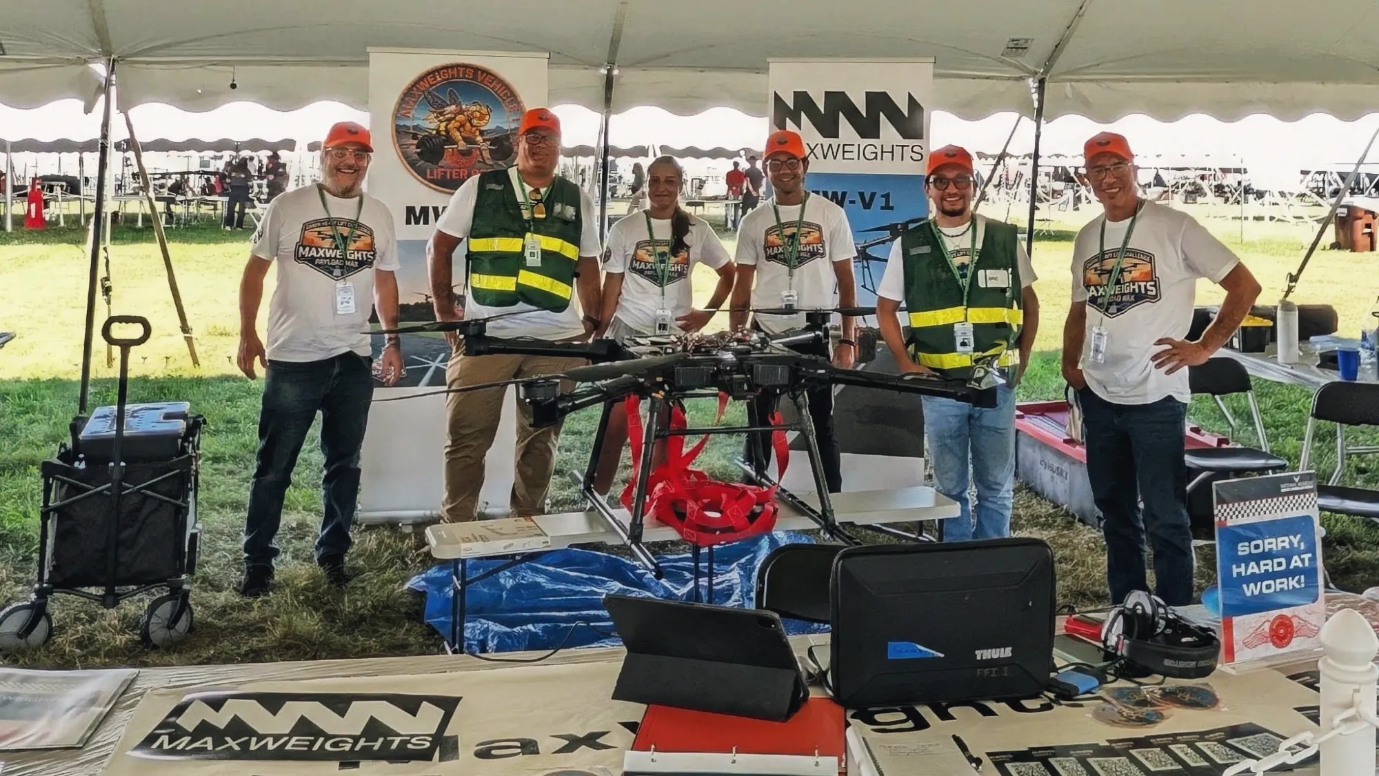 Six members of the Max Weights team standing behind the MW-V1 at their DARPA Lift Challenge booth, the red cargo harness rigged beneath the airframe.