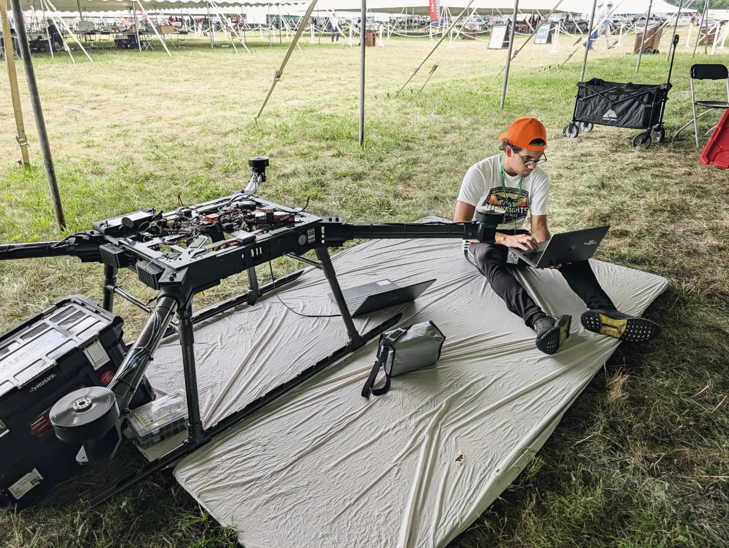 A team member sitting on a tarp beside the MW-V1 with a laptop open, reviewing flight data under the tent.