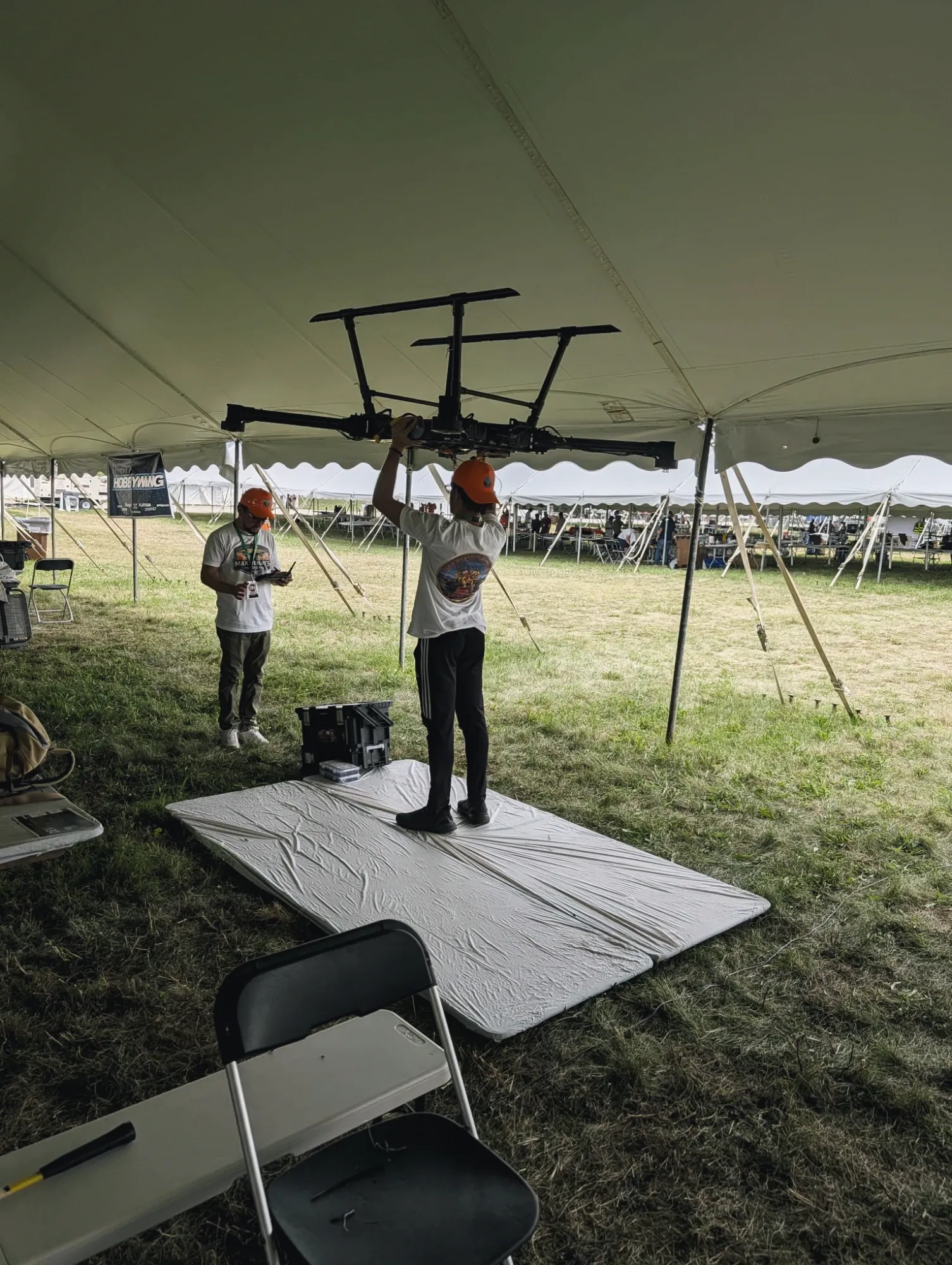 A team member lifting the bare MW-V1 airframe overhead under the competition tent while another checks a tablet.